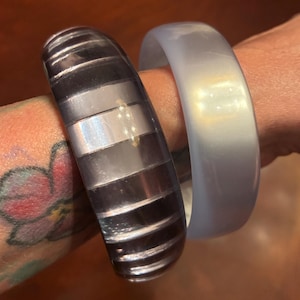 May include: Two bangles are shown. One is a silver and black striped design. The other is a solid, light blue bangle. The bangles are worn on a wrist. The background is a warm brown color.