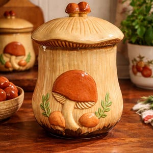 May include: A ceramic mushroom-shaped jar with a lid, featuring a brown mushroom design with green leaves. The jar is beige with a textured surface and sits on a wooden countertop. A matching jar is in the background.