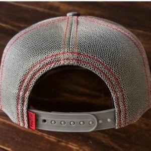 May include: Gray trucker hat with a mesh back and a snap closure. The hat features red stitching and a small red tag on the back. The hat is designed to be adjustable.