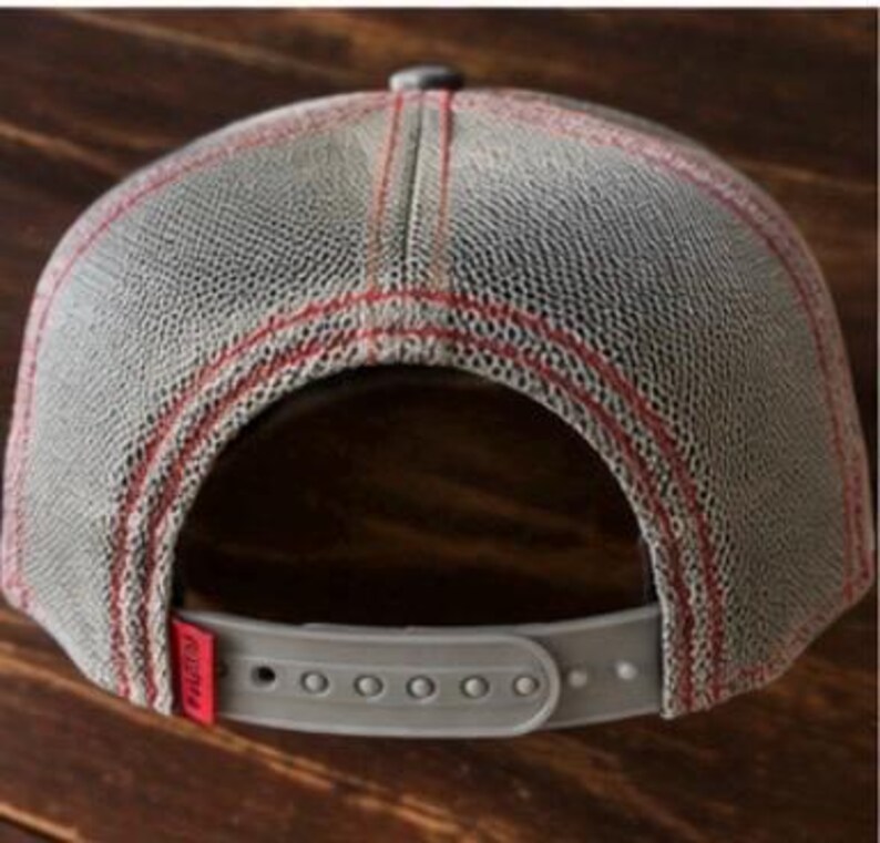 May include: Gray trucker hat with a mesh back and a snap closure. The hat features red stitching and a small red tag on the back. The hat is designed to be adjustable.