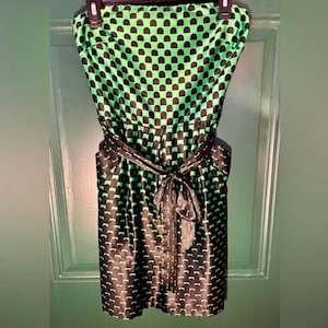 Puede incluir: Un vestido sin tirantes con un estampado negro y verde. El vestido presenta un patr&oacute;n repetitivo de formas negras sobre un fondo verde. Una faja negra est&aacute; atada a la cintura. El vestido est&aacute; colgado de una percha negra.
