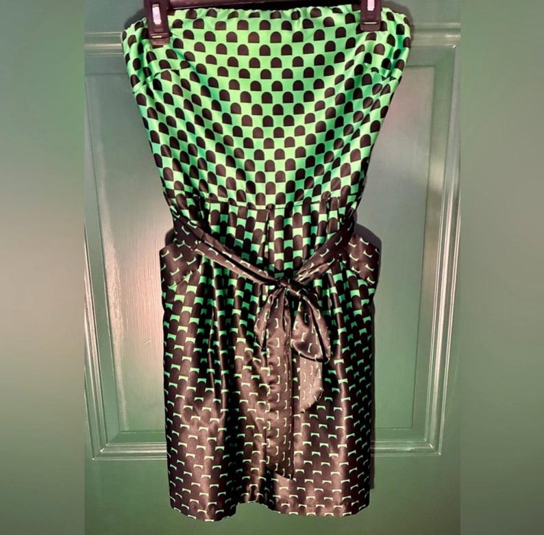 Puede incluir: Un vestido sin tirantes con un estampado negro y verde. El vestido presenta un patr&oacute;n repetitivo de formas negras sobre un fondo verde. Una faja negra est&aacute; atada a la cintura. El vestido est&aacute; colgado de una percha negra.