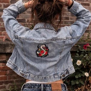 May include: A light blue denim jacket featuring a long-sleeved design with a collar and buttoned cuffs. The back of the jacket displays a central embroidered patch depicting a coiled snake surrounded by red roses and green leaves.