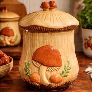 May include: A ceramic mushroom-shaped jar with a lid, featuring a detailed mushroom design in shades of brown and beige. The jar has a textured surface and is decorated with a large, raised mushroom and smaller mushrooms. The lid is topped with two small mushroom-shaped knobs.