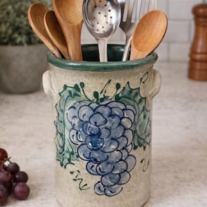 May include: A ceramic utensil holder with a hand-painted blue grape design and green accents. The holder contains wooden spoons, a slotted spoon, and a spatula. The holder has a green rim and small handles.