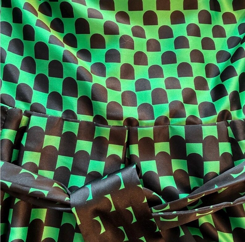 Puede incluir: Primer plano de una tela estampada verde y negra. El patr&oacute;n consiste en formas de semic&iacute;rculo negro dispuestas en una cuadr&iacute;cula sobre un fondo verde brillante. Un cintur&oacute;n de tela a juego es visible en la parte inferior, agregando un elemento de dise&ntilde;o cohesivo. La tela parece ser sat&eacute;n o seda.