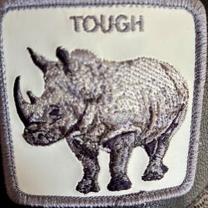 May include: A black and gray baseball cap featuring a patch with a detailed embroidered rhinoceros. The patch has the word "TOUGH" above the rhino. The cap has a leather-like texture and a patterned underside.