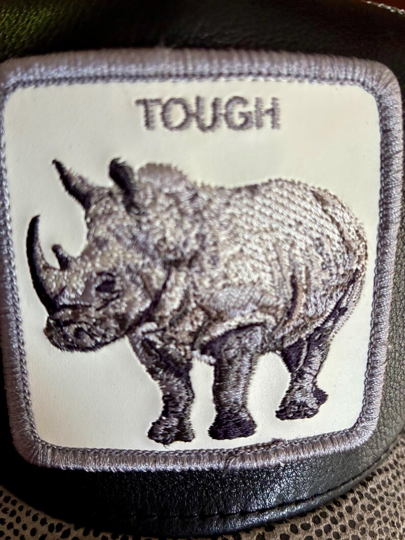 May include: A black and gray baseball cap featuring a patch with a detailed embroidered rhinoceros. The patch has the word "TOUGH" above the rhino. The cap has a leather-like texture and a patterned underside.