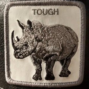 May include: Embroidered patch featuring a detailed rhino design in shades of gray. The word "TOUGH" is embroidered above the rhino. The patch has a white background with a gray border, suitable for jackets, bags, or other apparel.