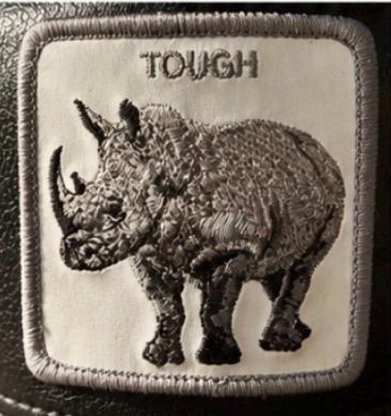 May include: Embroidered patch featuring a detailed rhino design in shades of gray. The word "TOUGH" is embroidered above the rhino. The patch has a white background with a gray border, suitable for jackets, bags, or other apparel.