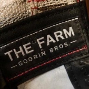 May include: Close-up of a black label with white text that reads "THE FARM - GOORIN BROS." The label is sewn onto a baseball cap with a mesh back. Red stitching is visible.