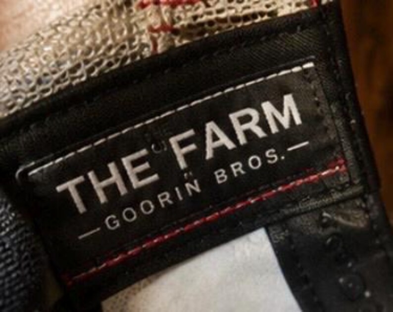May include: Close-up of a black label with white text that reads "THE FARM - GOORIN BROS." The label is sewn onto a baseball cap with a mesh back. Red stitching is visible.