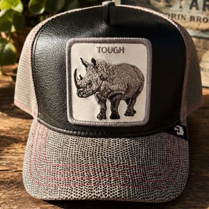 May include: A black and gray baseball cap with a rhino patch. The patch has the word "TOUGH" above a detailed illustration of a rhinoceros. The cap features a mesh back and a patterned brim with red stitching.