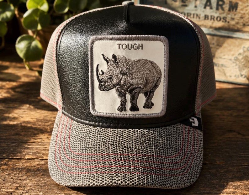 May include: A black and gray baseball cap with a rhino patch. The patch has the word "TOUGH" above a detailed illustration of a rhinoceros. The cap features a mesh back and a patterned brim with red stitching.