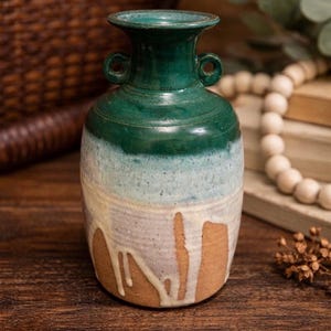 May include: A handcrafted ceramic vase with a two-tone glaze. The top is a deep green, transitioning to a light blue and cream color. The vase has two small handles and a textured surface, perfect for home decor.