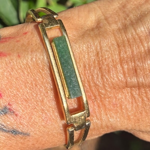 May include: A gold-toned metal bracelet featuring a rectangular link design. The central element is a long, narrow, dark green rectangular stone inlay set within a polished gold-colored frame. The bracelet consists of connected open metal links.