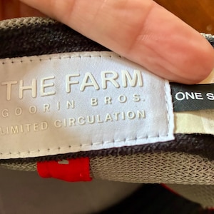 May include: A close-up of a baseball cap with a white label that reads "THE FARM GOORIN BROS. LIMITED CIRCULATION." A black tag reads "ONE SIZE." The cap has a red accent and a gray and black pattern.
