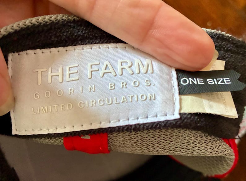 May include: A close-up of a baseball cap with a white label that reads "THE FARM GOORIN BROS. LIMITED CIRCULATION." A black tag reads "ONE SIZE." The cap has a red accent and a gray and black pattern.