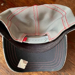 May include: A gray mesh baseball cap with red stitching and a red top. The underside of the brim is black with pink stitching. The cap has a white adjustable snap closure.