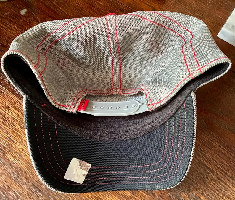May include: A gray mesh baseball cap with red stitching and a red top. The underside of the brim is black with pink stitching. The cap has a white adjustable snap closure.