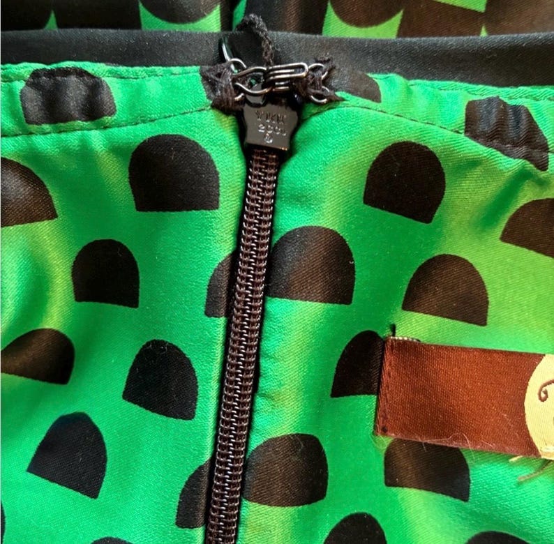 Puede incluir: Primer plano de una tela verde con un estampado de lunares negros. Una cremallera negra recorre verticalmente el centro. La tela forma parte de una prenda, con una etiqueta marr&oacute;n visible a la derecha.