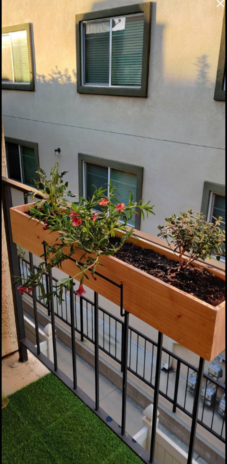 May include: A wooden planter box with a black metal bracket, hanging on a black metal railing. The planter box is filled with soil and has a variety of green plants and pink flowers growing in it.