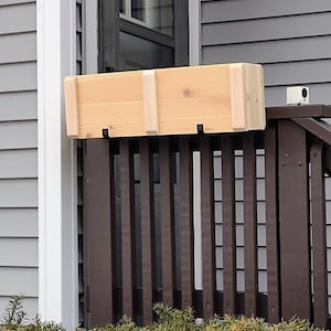 May include: A wooden planter box with a natural finish is mounted on a brown wooden railing. The planter box is attached to the railing with two black metal hooks.