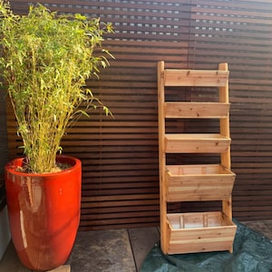 May include: A wooden plant stand with four shelves, made of natural wood. The stand is in front of a wooden slat wall and a potted plant.