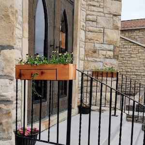 May include: Two wooden planter boxes with flowers are mounted on a black metal railing in front of a stone building with a large wooden door. The door has arched windows and is decorated with iron accents.