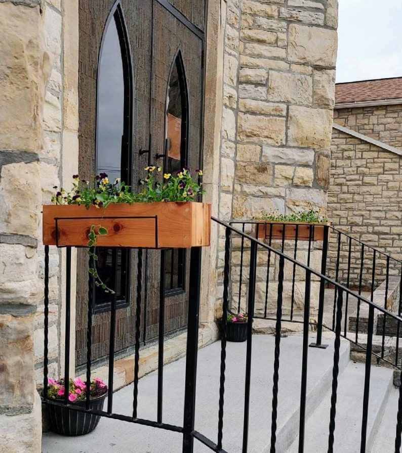 May include: Two wooden planter boxes with flowers are mounted on a black metal railing in front of a stone building with a large wooden door. The door has arched windows and is decorated with iron accents.