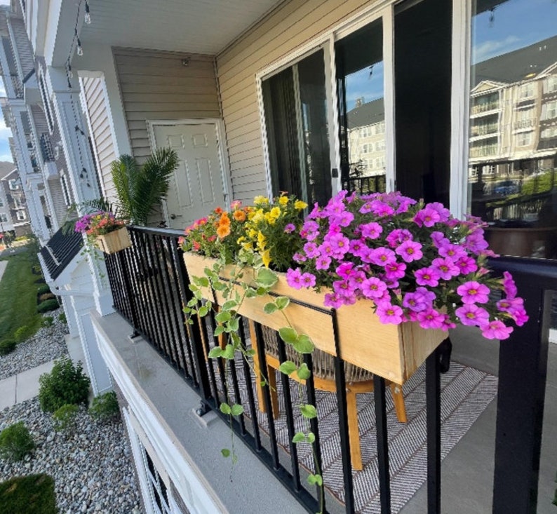 May include: A balcony with a black metal railing and a wooden planter box filled with pink and purple flowers. The planter box is mounted to the railing and is hanging over the edge of the balcony. The balcony is decorated with a black and white striped rug.