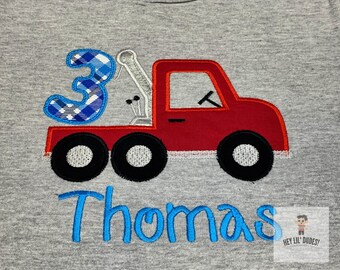 May include: A red toy truck with a blue and white checkered number 3 on the back. The name Thomas is embroidered in blue below the truck.
