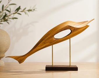 Può includere: Una scultura di pesce in legno stilizzata con una venatura liscia color marrone chiaro, caratterizzata da un ritaglio ovale centrale. La scultura è montata su due sottili aste metalliche verticali color ottone fissate a una base rettangolare in legno marrone scuro.