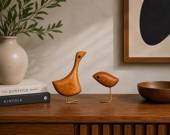 May include: Two wooden bird figurines with smooth, polished brown surfaces and dark circular eyes. Each bird features a slender, gold-toned metal wire leg and foot structure. The larger bird stands upright with a long neck, while the smaller bird is positioned in a forward-leaning posture.