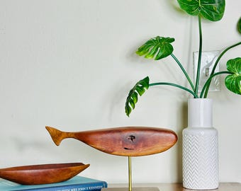 Può includere: Una scultura di balena in legno liscio intagliato a mano, montata su una sottile asta in ottone e una base rettangolare in legno scuro. Accanto si trova una piccola ciotola in legno curva appoggiata su un libro blu intitolato A TOUR OF THE WHITE HOUSE WITH MRS JOHN F KENNEDY.