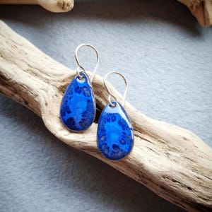 Puede incluir: Un par de pendientes azules en forma de lágrima con ganchos plateados. Los pendientes tienen un patrón veteado en tonos azules y se exhiben sobre un trozo de madera flotante sobre un fondo gris. Los pendientes son una joya.