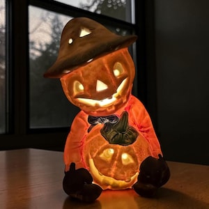 May include: A ceramic jack-o'-lantern figurine featuring a character wearing a wide-brimmed hat and holding a smaller pumpkin. The orange-toned piece is illuminated from within, casting a warm glow through the carved eyes, nose, and mouth.