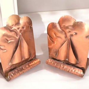 May include: A pair of copper bookends featuring a raised, embossed design of two stylized angel figures with wings. Each bookend has a smooth, reflective metallic finish and a sturdy, rectangular base with a textured, horizontal front edge.