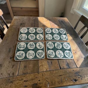 May include: A set of four rectangular placemats featuring a dark green and cream color scheme. Each placemat displays a grid of circular illustrations including animals like dogs, cows, and geese, as well as houses, hearts, and floral motifs.