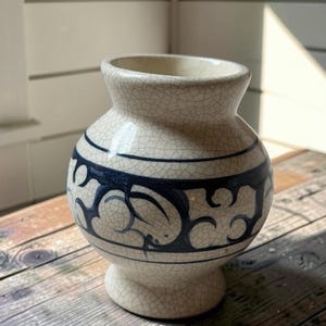 May include: A cream-coloured ceramic vase with a distinct crackle glaze finish. The vase features a rounded body, a flared rim, and a pedestal base. A dark blue hand-painted decorative band with swirling, abstract patterns encircles the centre.