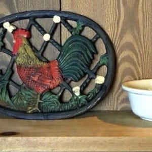 Könnte beinhalten: Ein gusseiserner Topfuntersetzer mit einem Hahn-Design in Rot, Grün und Gelb. Der Topfuntersetzer hat eine ovale Form mit einem dekorativen Gittermuster und steht auf einem Holzregal. Neben dem Topfuntersetzer steht eine weiße Schüssel.