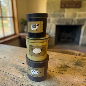May include: A stack of three cylindrical containers with lids. The top container is black with a single sheep illustration. The middle container is yellow with a sheep illustration. The bottom container is dark brown with three sheep illustrations.