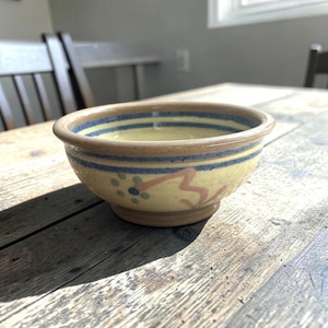 Pode incluir: Uma tigela de cerâmica redonda com base amarelo-claro, apresentando listras azuis horizontais ao redor da borda e um motivo floral pintado à mão em rosa e azul no exterior. A tigela tem um acabamento liso e vidrado e uma base circular elevada.