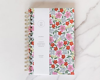 May include: A white journal with a floral pattern featuring pink, red, and blue flowers. The journal has a gold spiral binding and a white label with the text "EAST WEST studio" and "JOURNAL" along with the text "a journal for documenting your thoughts, dreams, and task lists" and "ruled pages 75 sheets".