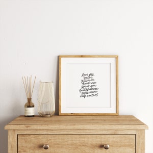 May include: A framed print with a white background and black text. The text reads "love joy, peace, patience, kindness, goodness, faithfulness, gentleness, self-control". The frame is a light wood color and is sitting on a wooden table.