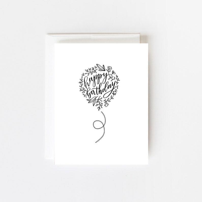 May include: A black and white greeting card with a balloon illustration. The balloon is surrounded by a wreath of flowers and leaves. The text inside the balloon reads "happy birthday".
