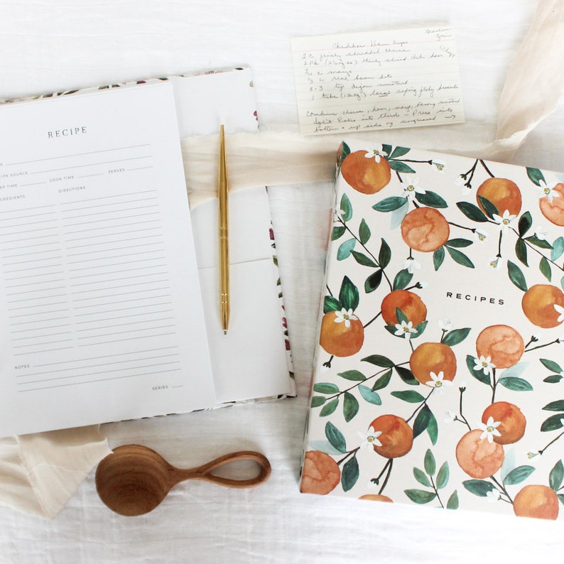 May include: A white recipe binder with a floral design of orange blossoms and leaves. The binder has the word "RECIPES" printed on the front.