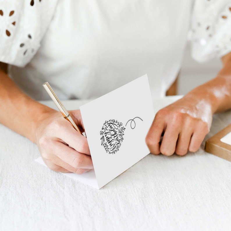 May include: A white card with a black floral wreath design that says "Happy Holidays" inside the wreath. A person is writing on the card with a gold pen.