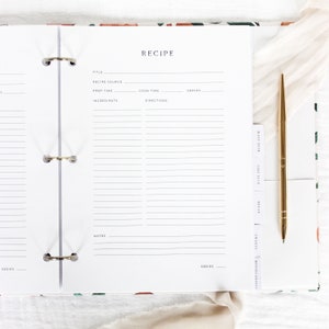 May include: A white recipe binder with a floral design on the cover. The binder has a gold ring mechanism and includes tabs for different recipe categories, such as main dish, side dish, and dessert.