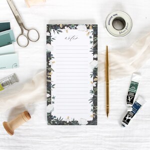 May include: A white notepad with a black floral border and the word "notes" written in black cursive. The notepad is on a white surface with other craft supplies.